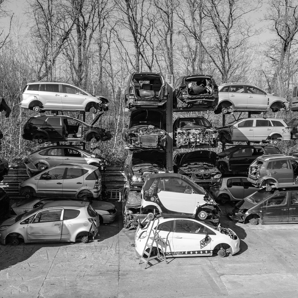 Schwarz-Weiß-Foto vom Autoplatz bei Jungkurth. In hohen Hochregalen stapeln sich zahlreiche Karosserien von Altfahrzeugen vor einer Waldkulisse, wobei einzelne Autos in einem auffälligen Türkis hervorgehoben sind.