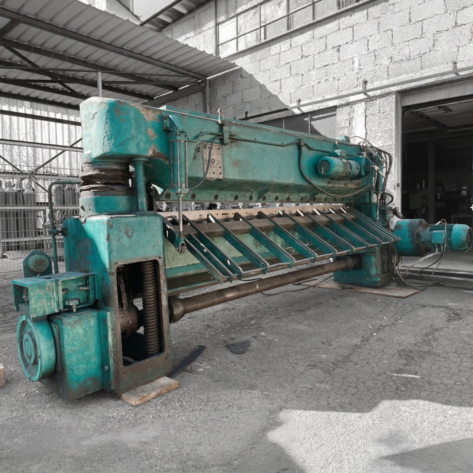 Eine großformatige, türkisgrüne Blechschere von Jungkurth, die als farblicher Akzent vor einem in Schwarz-Weiß gehaltenen Werkstattgebäude steht. Die massive Maschine zeigt deutliche Gebrauchsspuren, freiliegende mechanische Wellen und Elektromotoren auf einem asphaltierten Außenbereich.