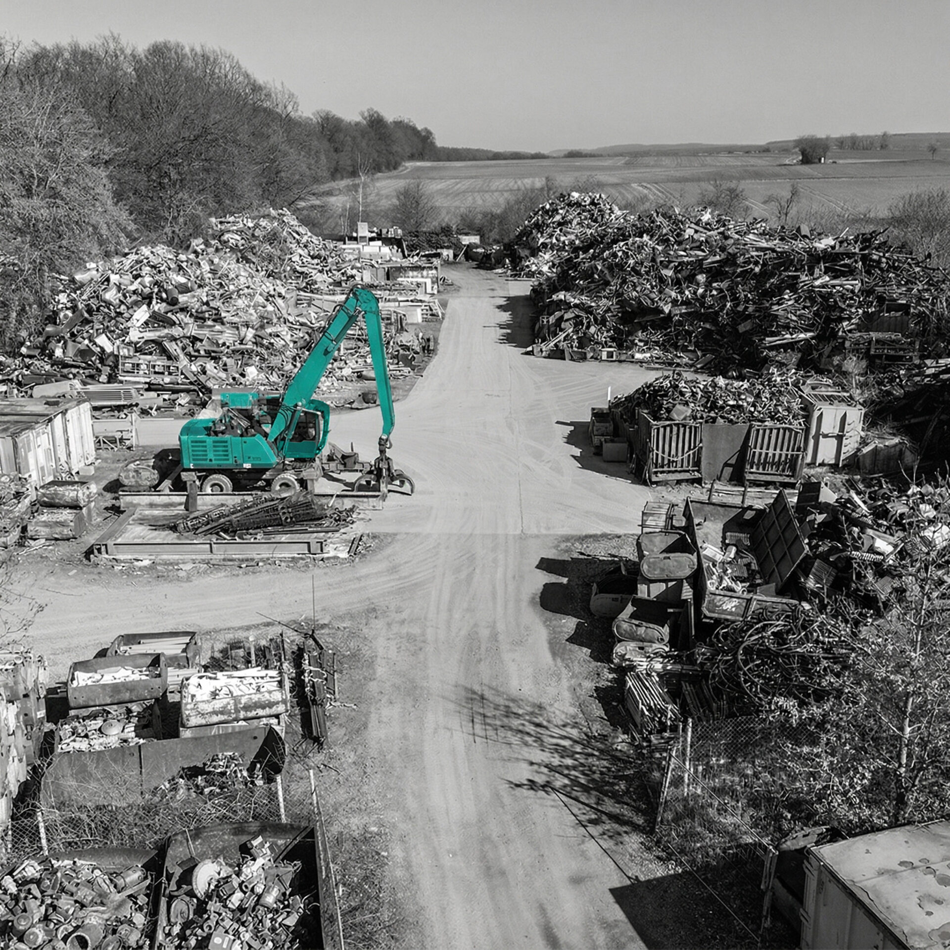 Eine Luftaufnahme des Schrottplatzes von Jungkurth in Schwarz-Weiß, auf der ein türkisfarbener Umschlagbagger als einziger Farbakzent hervorsticht. Der Bagger steht auf einem zentralen Fahrweg zwischen hohen Bergen aus sortiertem Metallschrott, Containern und Gitterboxen vor einer ländlichen Kulisse.